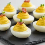 Close-up of several classic deviled eggs arranged on a white platter. Each egg half features a swirl of creamy yellow filling, garnished with red paprika and finely chopped green herbs.