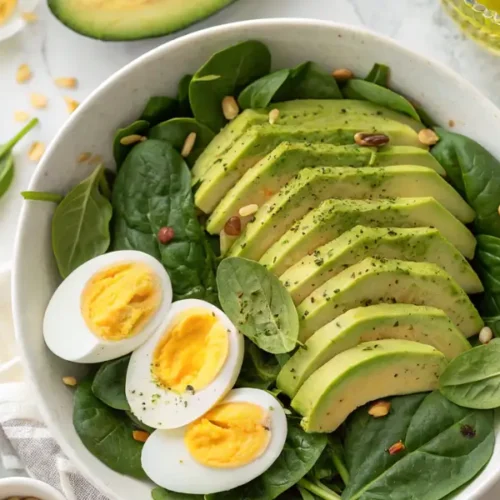 Healthy avocado egg and spinach salad in white bowl with sliced avocado and hard-boiled eggs on fresh spinach leaves