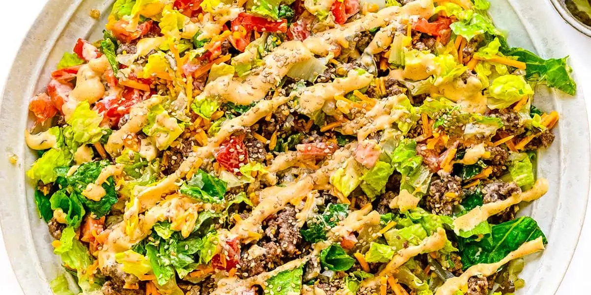 Low-carb Big Mac salad recipe in a bowl with ground beef, special sauce, cheese, pickles, and sesame seeds on a wood table